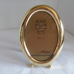 May include: An oval-shaped solid brass photo frame with a polished gold-tone finish. The frame features a clear glass front with printed text reading SOLID BRASS Photo Frame hand polished lacquer coated 3 1/2 x 5 and the brand name Allegro.