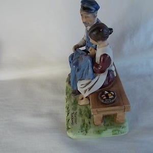 May include: A porcelain figurine of a man and a girl sitting on a bench. The man is wearing a blue and white outfit and a hat. The girl is wearing a red and white dress. The bench is brown and has a basket of fruit on it. The figurine is signed "Norman Rockwell".