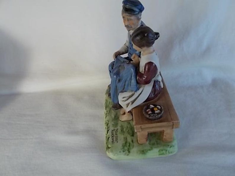 May include: A porcelain figurine of a man and a girl sitting on a bench. The man is wearing a blue and white outfit and a hat. The girl is wearing a red and white dress. The bench is brown and has a basket of fruit on it. The figurine is signed "Norman Rockwell".