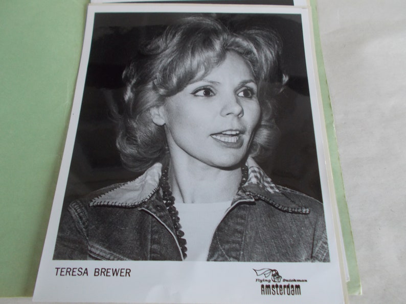 May include: Black and white photo of Teresa Brewer, a singer, wearing a denim jacket and a checkered shirt. The photo is signed "Teresa Brewer" and has a logo for "Flying Dutchman Amsterdam" at the bottom.