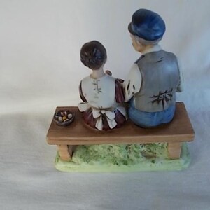May include: A porcelain figurine of a man and a woman sitting on a wooden bench. The man is wearing a blue hat and a gray vest. The woman is wearing a white dress with a brown apron. They are both looking to the right. There is a small basket of fruit in front of them.