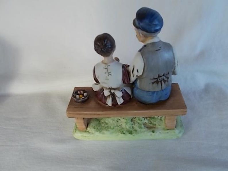 May include: A porcelain figurine of a man and a woman sitting on a wooden bench. The man is wearing a blue hat and a gray vest. The woman is wearing a white dress with a brown apron. They are both looking to the right. There is a small basket of fruit in front of them.