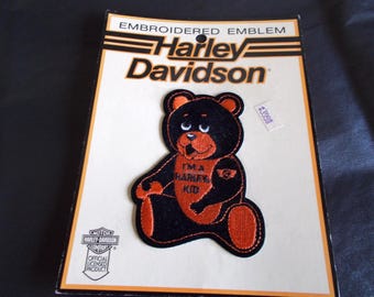 Puede incluir: Parche bordado de Harley-Davidson con un oso de peluche negro y naranja con el texto "I'M A HARLEY KID". El parche está sobre un fondo blanco con el logotipo de Harley-Davidson e información oficial del producto con licencia.