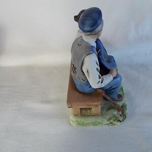 May include: A porcelain figurine of a man sitting on a bench with a cane. He is wearing a blue hat, a gray vest, and blue pants. The bench is brown and has a green base.