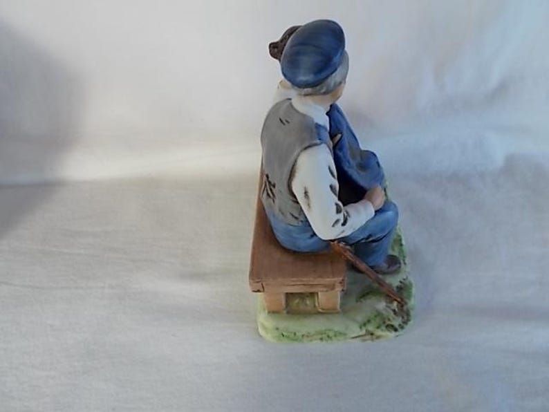 May include: A porcelain figurine of a man sitting on a bench with a cane. He is wearing a blue hat, a gray vest, and blue pants. The bench is brown and has a green base.