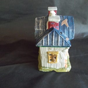 Może przedstawiać: Ceramiczny miniaturowy domek z niebieskim dachem, białymi ścianami i czerwonym ceglanym kominem. Dom ma małe okno z drewnianymi listwami i stoi na zielonej podstawie. Dach ma wygląd zniszczony.