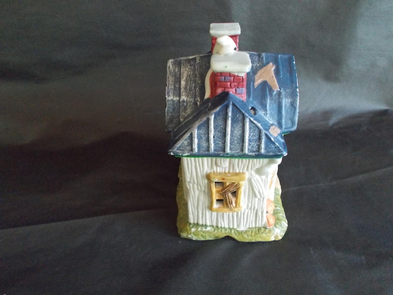 Może przedstawiać: Ceramiczny miniaturowy domek z niebieskim dachem, białymi ścianami i czerwonym ceglanym kominem. Dom ma małe okno z drewnianymi listwami i stoi na zielonej podstawie. Dach ma wygląd zniszczony.