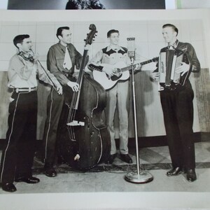 May include: A black and white photo of a band playing music. The band consists of four men, one playing a fiddle, one playing a bass, one playing a guitar, and one playing an accordion.