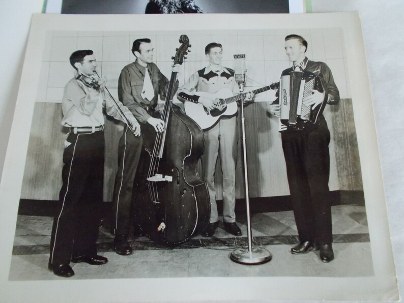 May include: A black and white photo of a band playing music. The band consists of four men, one playing a fiddle, one playing a bass, one playing a guitar, and one playing an accordion.