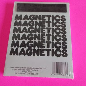 May include: A rectangular notepad with a light gray cover featuring the word MAGNETICS printed in black six times in a stacked pattern. A black magnetic strip is attached to the top. The bottom includes a barcode and copyright text.