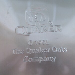 Op de afbeelding: Een zilveren metalen Quaker Oats Company-logo met de tekst "Quaker" en een gestileerde afbeelding van een man met een hoed. De tekst "&copy; 1991 The Quaker Oats Company" staat onder het logo.