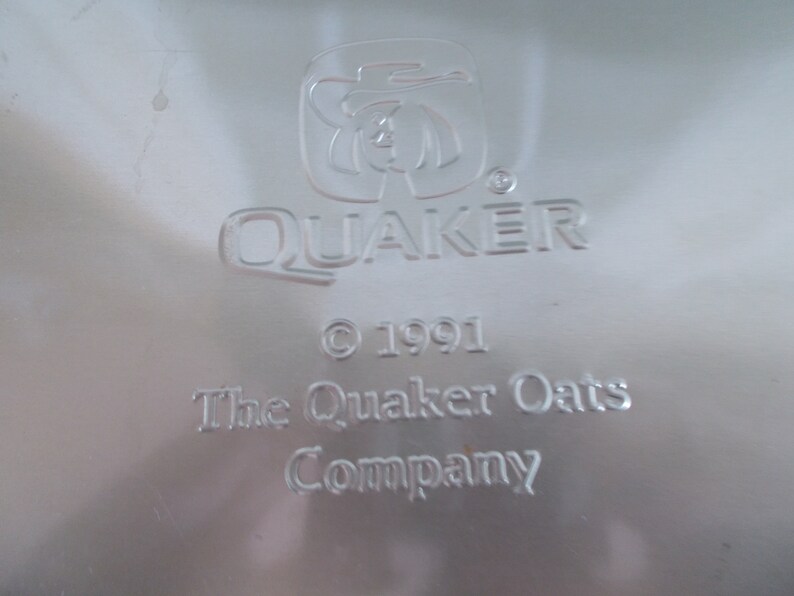 Op de afbeelding: Een zilveren metalen Quaker Oats Company-logo met de tekst "Quaker" en een gestileerde afbeelding van een man met een hoed. De tekst "&copy; 1991 The Quaker Oats Company" staat onder het logo.