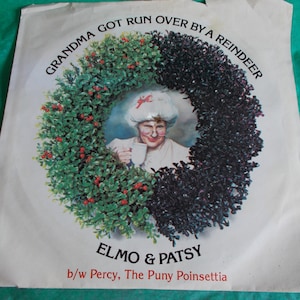May include: A white vinyl record with a green and black wreath on the cover. The wreath surrounds a picture of a woman wearing a white hat and holding a cup. The text on the record reads "Grandma Got Run Over By A Reindeer" and "Elmo & Patsy b/w Percy, The Puny Poinsettia".
