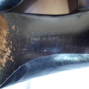 May include: Close-up of the sole of a black leather shoe. The sole shows wear and tear, with visible scuff marks and discoloration. The text "CAMPER" and "MADE IN SPAIN" are embossed on the sole.