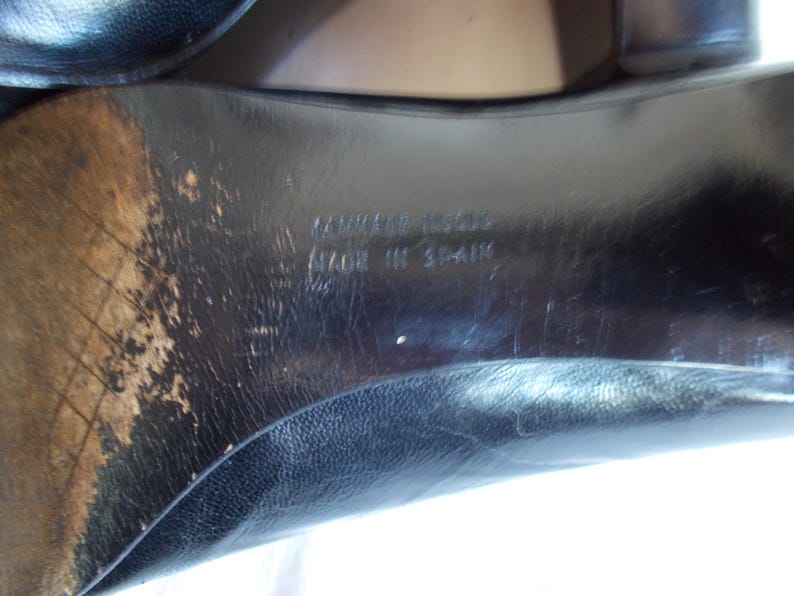 May include: Close-up of the sole of a black leather shoe. The sole shows wear and tear, with visible scuff marks and discoloration. The text "CAMPER" and "MADE IN SPAIN" are embossed on the sole.