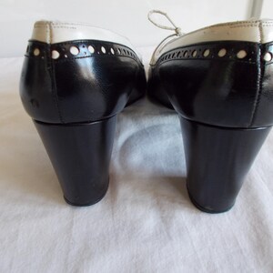 May include: Pair of black and white oxford-style shoes with a low, black heel. The shoes have a glossy finish with white detailing around the top and decorative perforations. The laces are a light cream color.