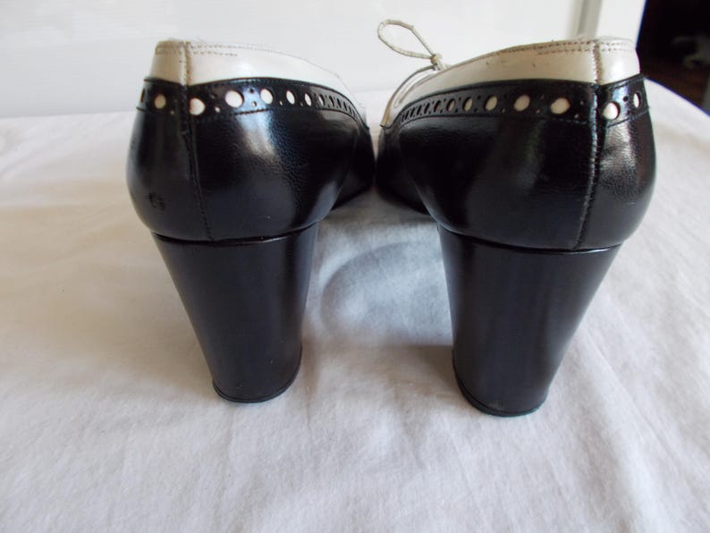 May include: Pair of black and white oxford-style shoes with a low, black heel. The shoes have a glossy finish with white detailing around the top and decorative perforations. The laces are a light cream color.