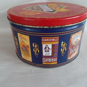 Op de afbeelding: Een ronde blauwe en rode blikken doos met het Quaker Oats logo op de deksel. De doos heeft een blauwe achtergrond met afbeeldingen van verschillende Quaker Oats producten, waaronder "Columbian Rolled White Oats", "Quaker Mills Oatmeal" en "Akron Oatmeal".