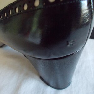 May include: Close-up of a black and cream-colored shoe. The shoe features a glossy black heel and a cream-colored upper with decorative perforations. The shoe has a classic, vintage style.