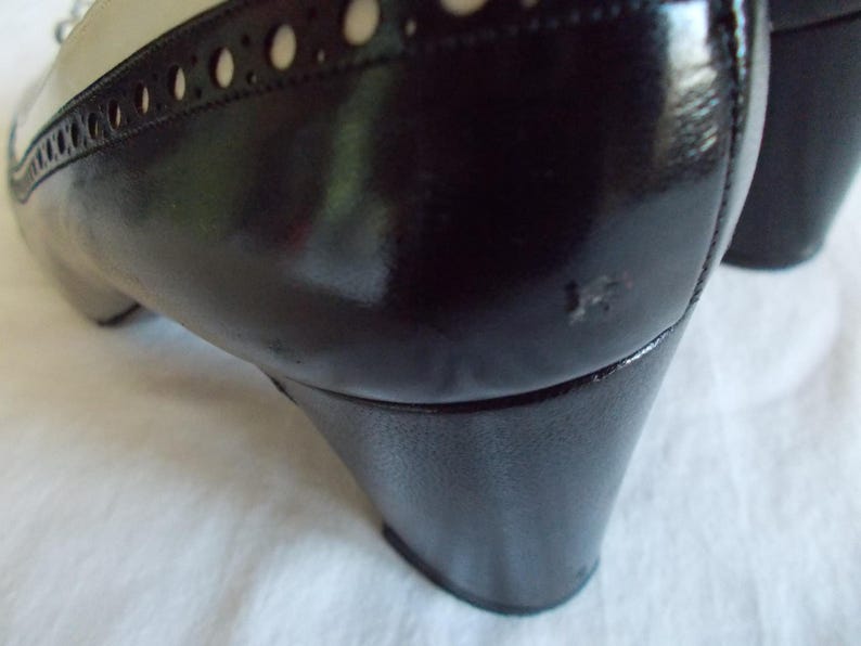 May include: Close-up of a black and cream-colored shoe. The shoe features a glossy black heel and a cream-colored upper with decorative perforations. The shoe has a classic, vintage style.