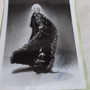 May include: Black and white photograph of a woman wearing a long black coat with a fur trim. She is standing in a studio setting and has a large feathered boa draped around her shoulders. The woman is smiling and looking at the camera. The photograph is signed by the woman in blue ink.