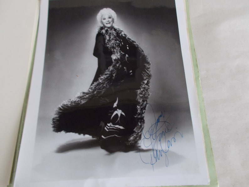 May include: Black and white photograph of a woman wearing a long black coat with a fur trim. She is standing in a studio setting and has a large feathered boa draped around her shoulders. The woman is smiling and looking at the camera. The photograph is signed by the woman in blue ink.