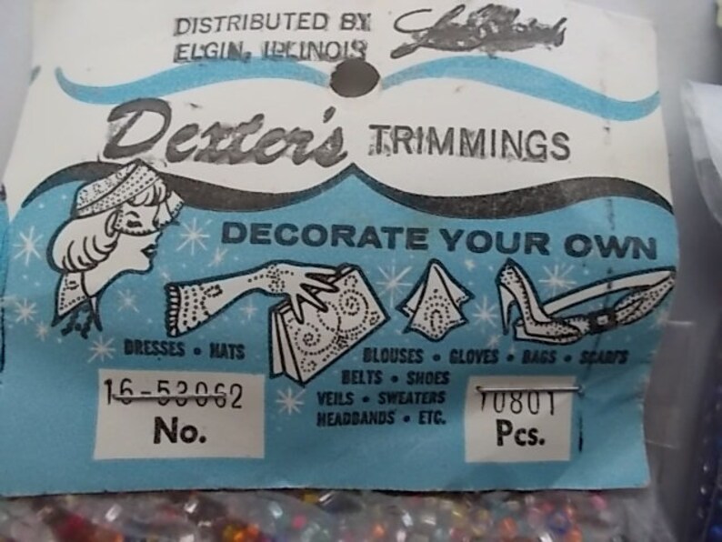 May include: Vintage Dexter's Trimmings label with a light blue and white design. The label features illustrations of dresses, hats, gloves, and shoes. Text includes "Decorate Your Own" and a list of items. The label also has a number and quantity.