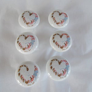May include: Six white ceramic cabinet knobs with a heart-shaped design. Each knob features a brown rope-like border with pink and blue floral accents. The text "HOL 1983" is printed on the bottom of each knob.