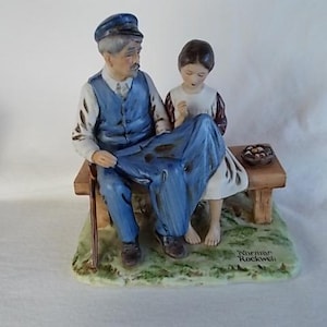 May include: A porcelain figurine of an older man with a blue cap and a young girl sitting on a wooden bench. The man is wearing a blue vest and blue pants. The girl is wearing a white dress with a red and white patterned bodice. The figurine is signed "Norman Rockwell".