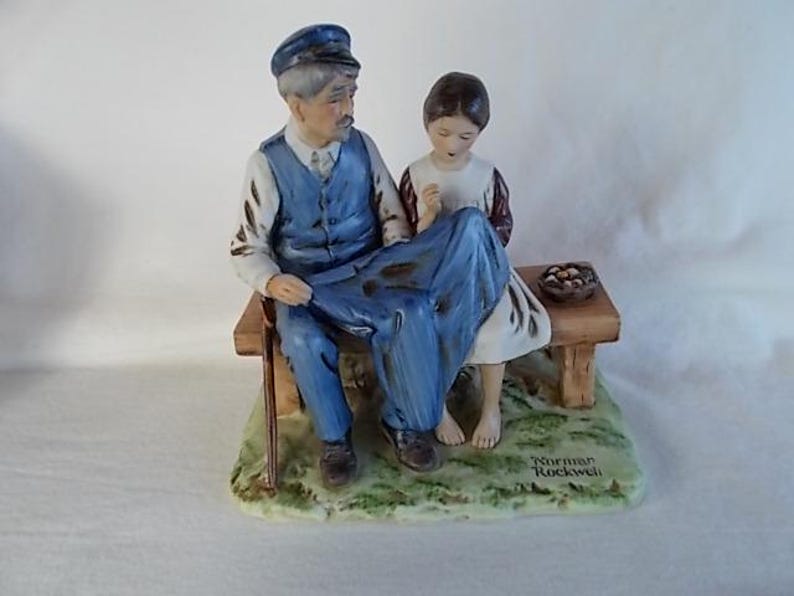 May include: A porcelain figurine of an older man with a blue cap and a young girl sitting on a wooden bench. The man is wearing a blue vest and blue pants. The girl is wearing a white dress with a red and white patterned bodice. The figurine is signed "Norman Rockwell".