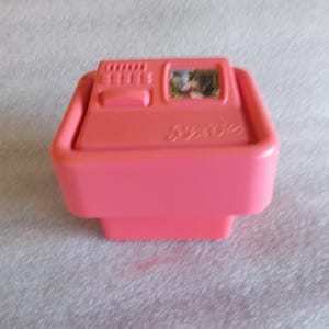 May include: A pink plastic toy cash register with a small picture on top. The register has a button and a series of small rectangular shapes. The word "Barbie" is printed on the front.
