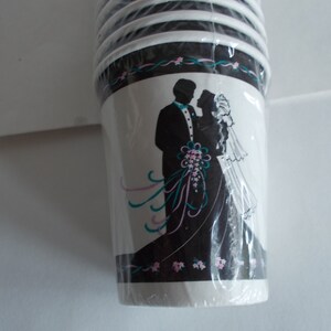 May include: A stack of white paper cups with a black and white silhouette of a bride and groom on the front. The cups have a teal and pink floral design around the top.