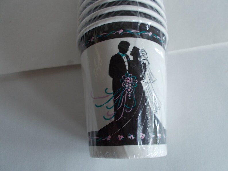 May include: A stack of white paper cups with a black and white silhouette of a bride and groom on the front. The cups have a teal and pink floral design around the top.