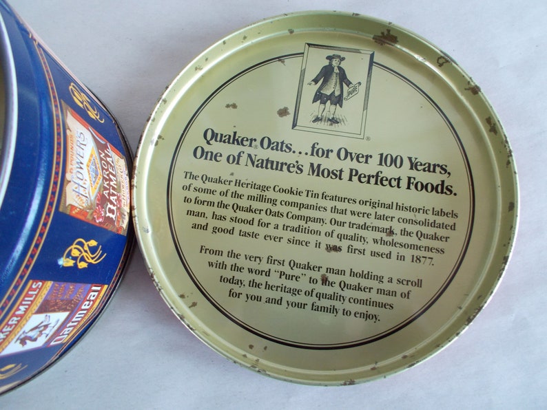 Op de afbeelding: Een vintage Quaker Oats koekjestrommel met een geel deksel en een zwart-witte illustratie van een man die een rol papier vasthoudt. Het deksel heeft de tekst "Quaker Oats...for Over 100 Years, One of Nature's Most Perfect Foods."