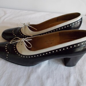 May include: A pair of black and white oxford-style heels. The shoes have a rounded toe, a cream-colored vamp, and a black body with white stitching. The heels are approximately 2.5 inches high.