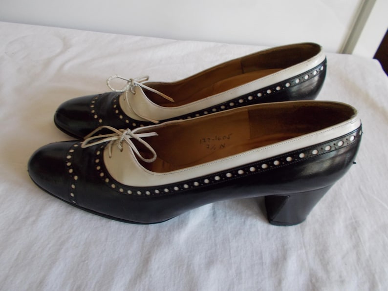 May include: A pair of black and white oxford-style heels. The shoes have a rounded toe, a cream-colored vamp, and a black body with white stitching. The heels are approximately 2.5 inches high.