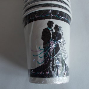 May include: A stack of white paper cups with a black and white silhouette of a bride and groom on the front. The bride is wearing a white dress and the groom is wearing a tuxedo. The cups have a black and white design with pink and teal accents.