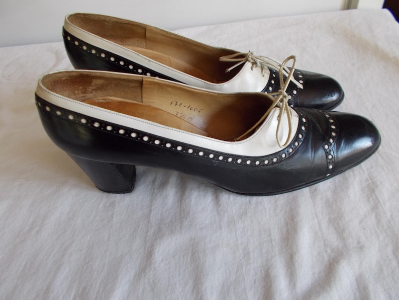 May include: A pair of vintage black and white leather shoes with a low heel. The shoes have a classic design with a white accent on the top and a lace-up closure. The shoes are in good condition.