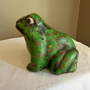 May include: A ceramic frog figurine in shades of green and brown. The frog has a textured surface and detailed features, including visible eyes. The figurine is a decorative item, likely for home decor or a collector's piece.