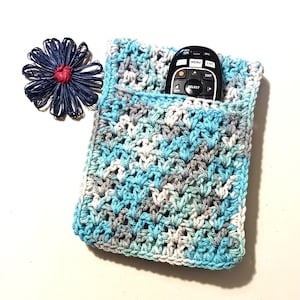 May include: Crocheted remote control holder in shades of blue, gray, and white, with a remote inside. A blue and red decorative flower is to the left of the holder. The holder is approximately 7 inches tall and 5 inches wide.