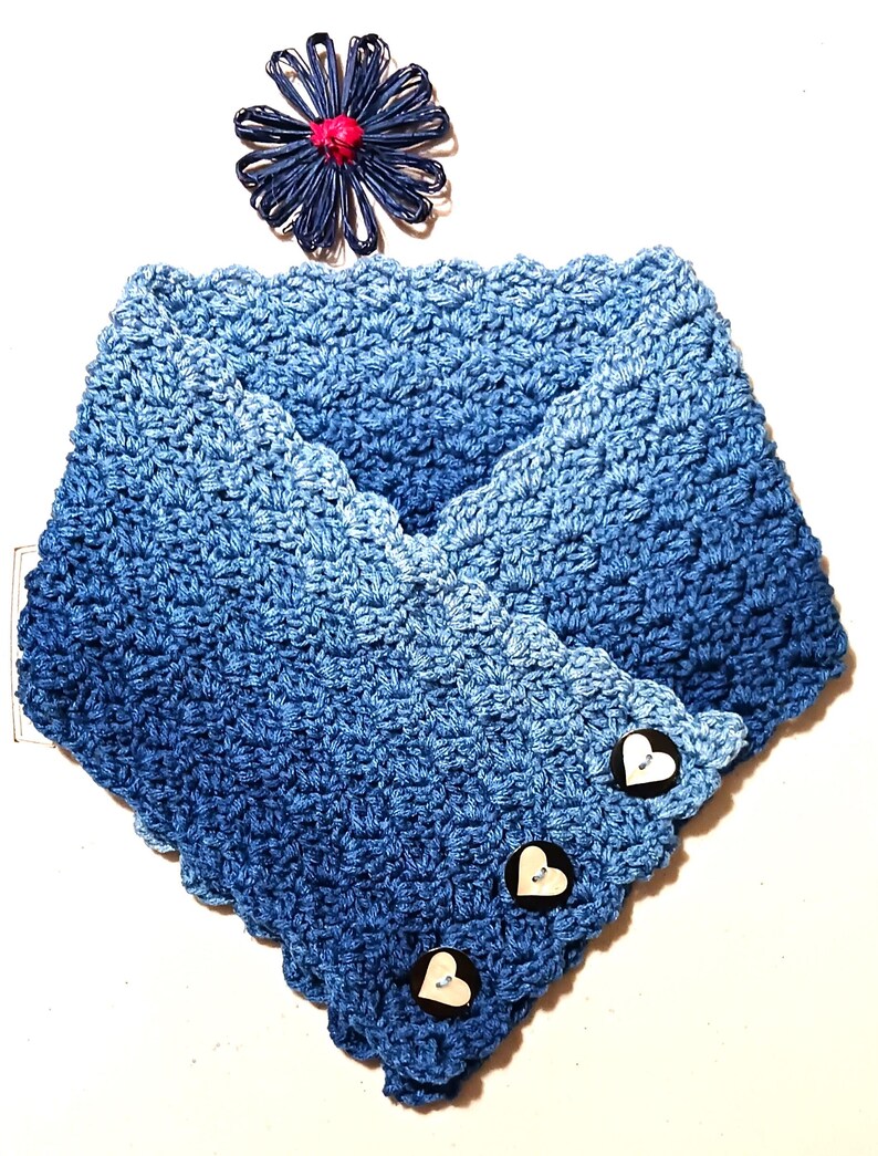 May include: A blue crocheted cowl with a gradient effect, transitioning from light to dark shades. The cowl features three heart-shaped buttons and a decorative blue flower. This handmade accessory is perfect for adding a touch of handmade charm to any outfit.