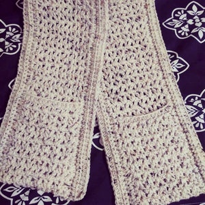 Pode incluir: Uma echarpe de croch&ecirc; com dois bolsos, feita com fio bege com manchas marrons e brancas. A echarpe est&aacute; dobrada ao meio e colocada sobre um tecido estampado em preto e branco.