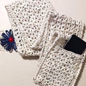 Pode incluir: Um bolso de telefone de croch&ecirc; branco com um smartphone preto dentro. O bolso &eacute; feito com um fio texturizado e tem uma apar&ecirc;ncia ligeiramente salpicada.