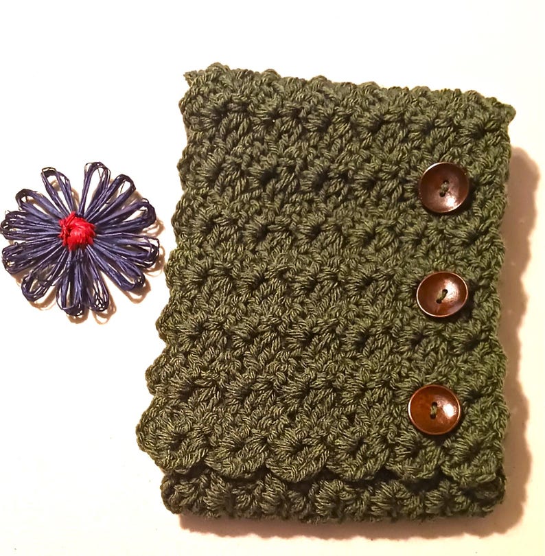 May include: An olive green crocheted sleeve with three brown buttons. A blue and red decorative flower is to the left of the sleeve. The sleeve has a textured pattern and a scalloped edge.