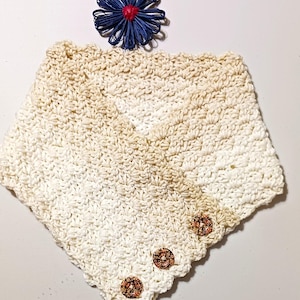 May include: Cream-coloured crocheted cowl with three decorative buttons. The cowl has a V-neck design and a textured stitch pattern. A blue and red decorative flower is placed above the cowl.