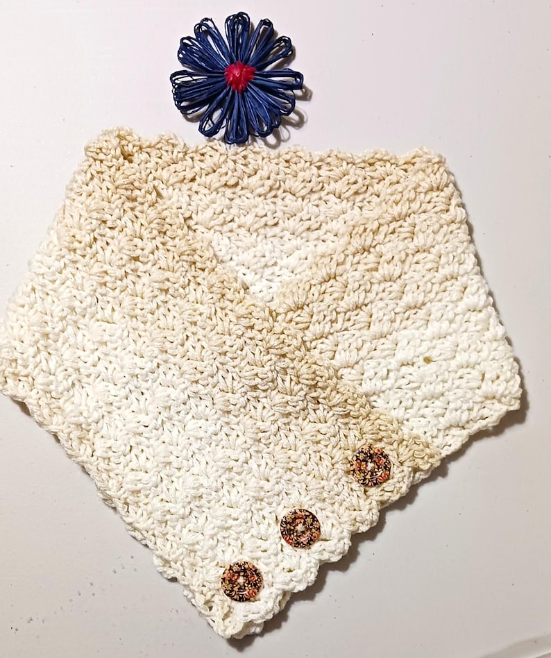 May include: Cream-coloured crocheted cowl with three decorative buttons. The cowl has a V-neck design and a textured stitch pattern. A blue and red decorative flower is placed above the cowl.