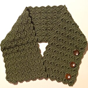 May include: An olive green crocheted cowl with a scalloped edge and three brown buttons. The neck warmer is designed to be worn around the neck and is made of a textured yarn.