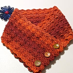 May include: A crocheted cowl in shades of orange and burgundy, with a button closure. The cowl has a V-neck design and is adorned with three wooden buttons. A blue decorative flower is placed to the left of the cowl.
