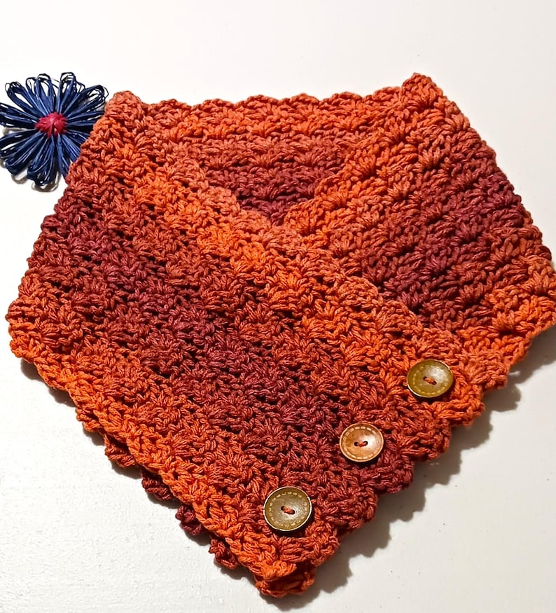 May include: A crocheted cowl in shades of orange and burgundy, with a button closure. The cowl has a V-neck design and is adorned with three wooden buttons. A blue decorative flower is placed to the left of the cowl.