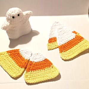 May include: A white crocheted ghost figure with two black eyes positioned next to three triangular crocheted candy corn pieces. Each candy corn features horizontal stripes of white, orange, and yellow yarn, creating a textured, layered appearance.